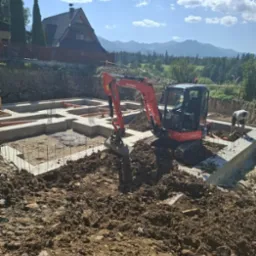 Fundamenty domu z małą koparką Kubota w akcji, malownicze góry w tle. Widoczne zbrojenia i szalunki. Prace ziemne w toku, słoneczny dzień.