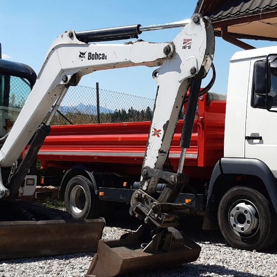 Mini koparka Bobcat z łyżką na żwirowym podłożu, w tle czerwona wywrotka i ogrodzenie z siatki. Sprzęt budowlany w trakcie prac ziemnych.