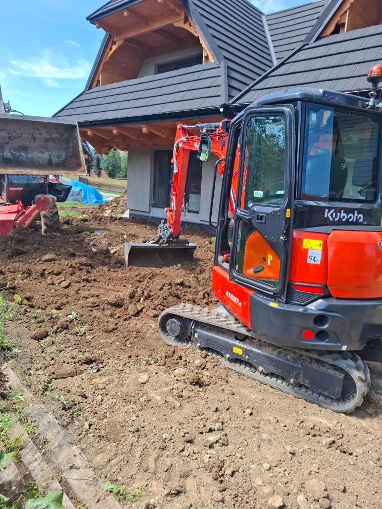 Pomarańczowa mini koparka Kubota KX037-4 na placu budowy przed domem z ciemnym dachem. W tle druga koparka i rozkopana ziemia.