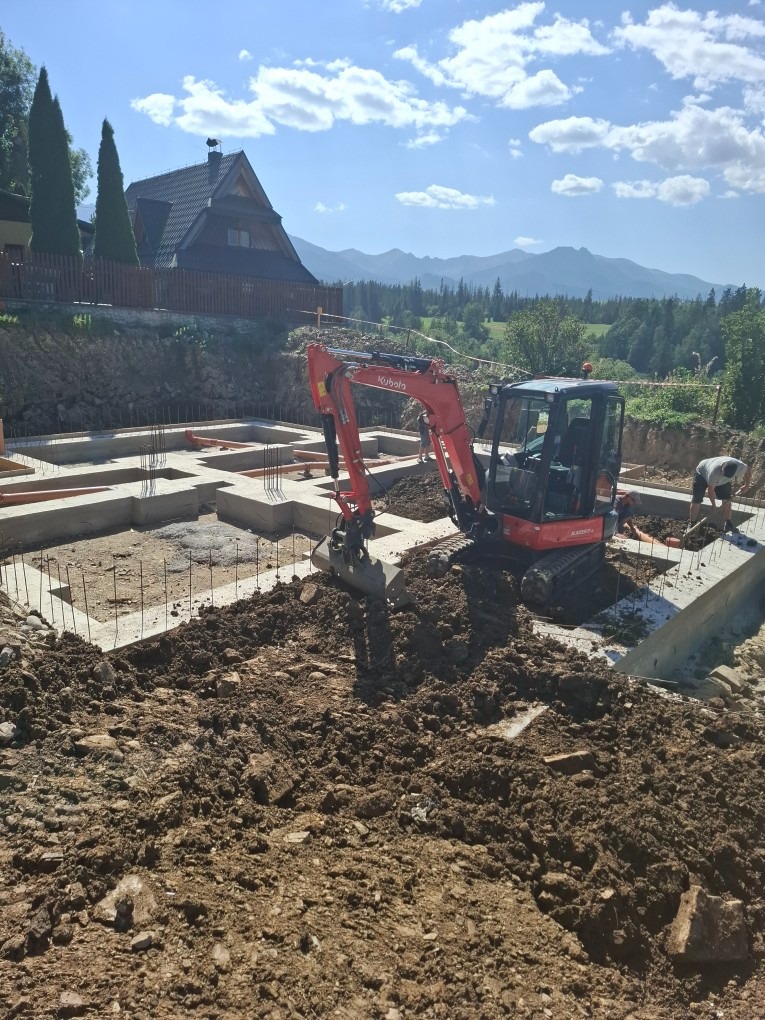 Fundamenty domu z małą koparką Kubota w akcji, malownicze góry w tle. Widoczne zbrojenia i szalunki. Prace ziemne w toku, słoneczny dzień.