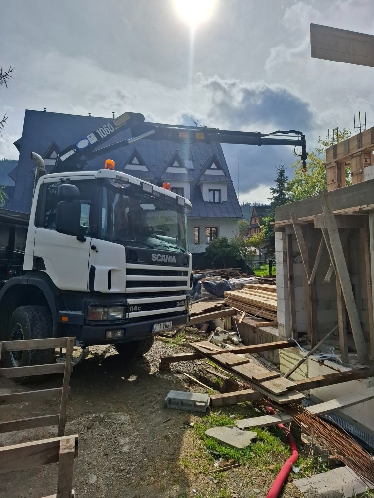 Ciężarówka Scania z dźwigiem na placu budowy, w tle dom z szarym dachem. Widoczne drewniane elementy konstrukcji i zbrojenia. Słoneczny dzień.
