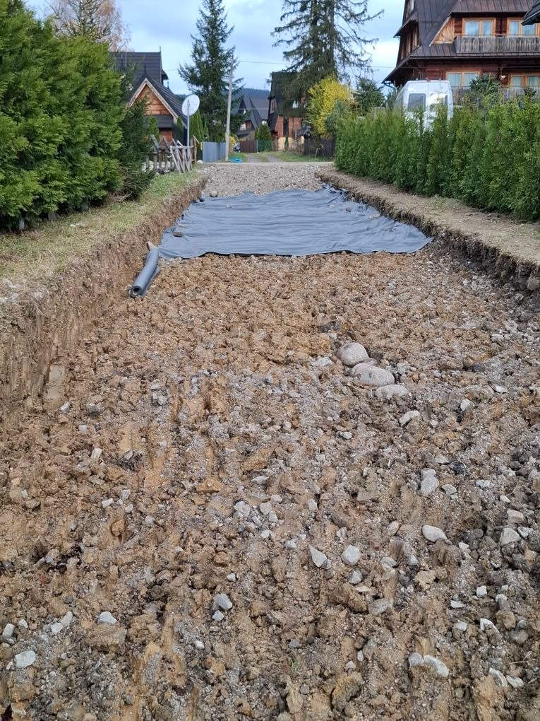 Wykop pod drogę z widoczną rurą drenażową i szarą geowłókniną. W tle zabudowa jednorodzinna i drzewa.