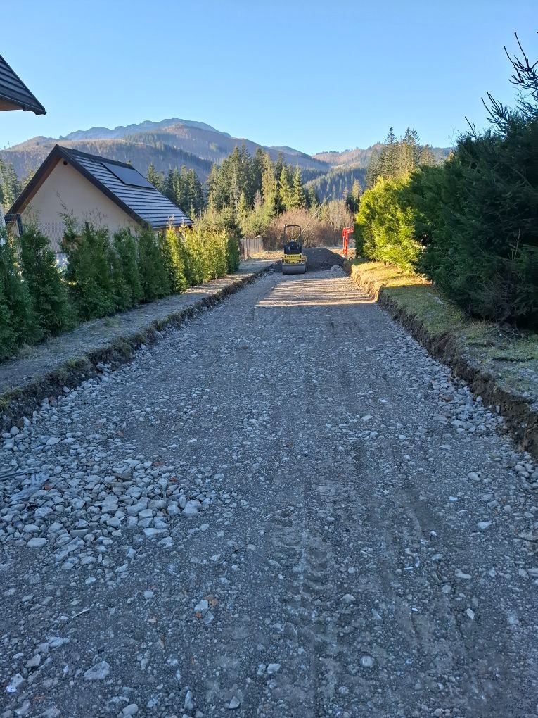 Droga z tłucznia w trakcie utwardzania z widokiem na góry w tle i małą maszyną budowlaną, otoczona zielenią i częścią domu po lewej stronie.