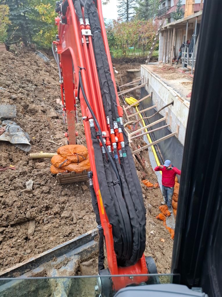 Czerwona koparka Kubota w trakcie wykopu pod fundamenty, widok z kabiny, pracownik w czerwonej bluzie nadzoruje prace. Żółte rury w wykopie, zabezpieczenia.