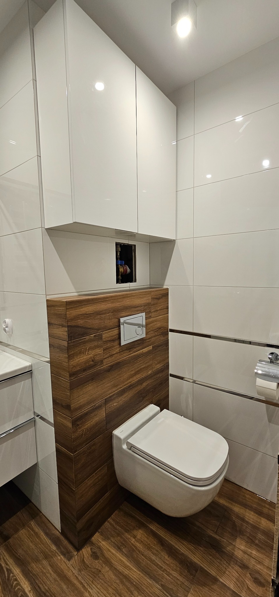 Nowoczesna toaleta podwieszana z drewnianą obudową stelaża, białe płytki z metalowymi listwami i białe szafki wiszące w wykończonej łazience.