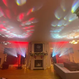 Eleganckie wnętrze z kominkiem, oświetlone dynamicznymi światłami scenicznymi w kolorach czerwieni i błękitu, tworzącymi wzory na suficie. Dekoracje świąteczne.