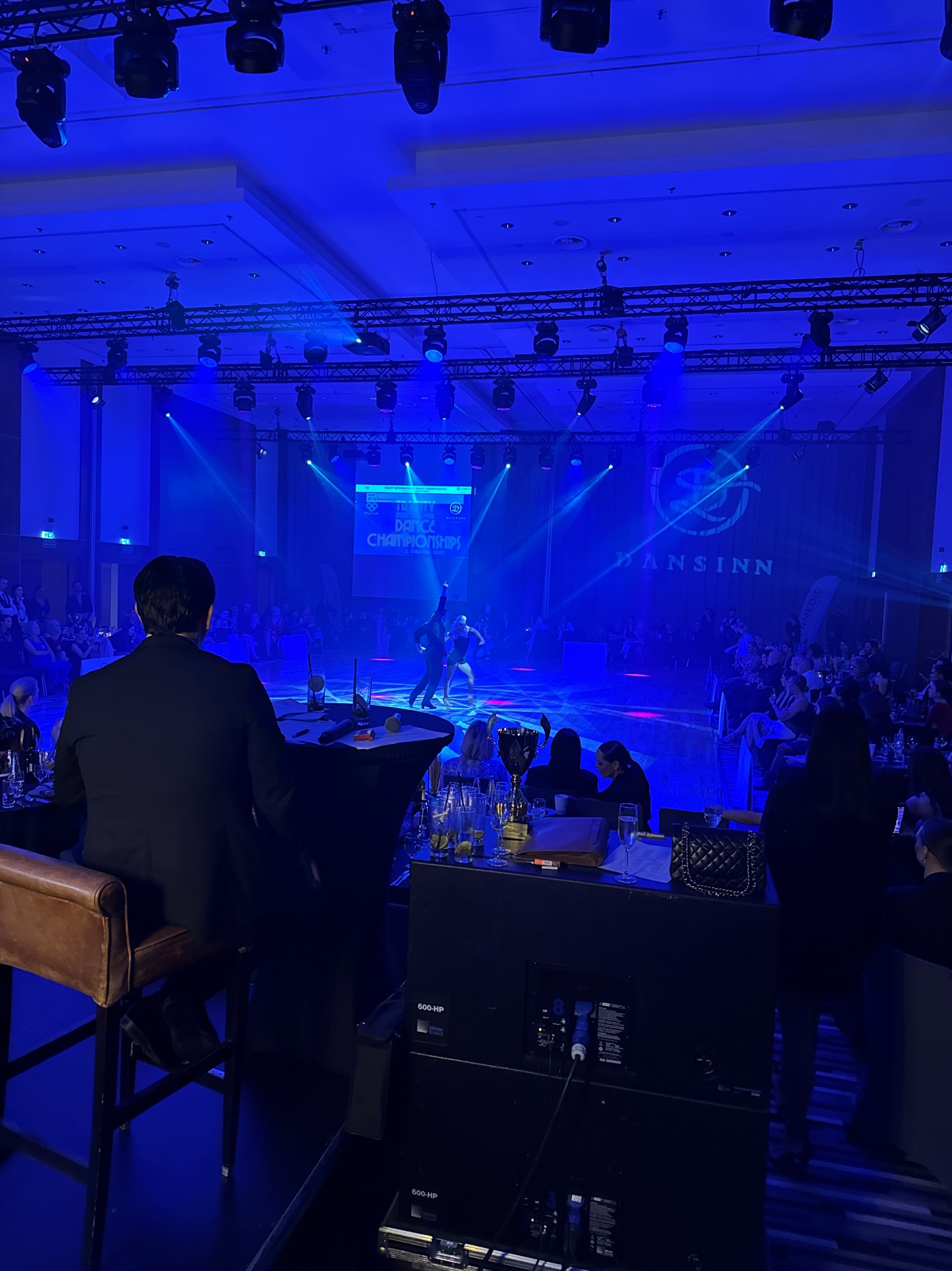 Widok zza kulis na taneczny turniej: parkiet skąpany w niebieskim świetle, tancerze w ruchu, publiczność przy stolikach, sprzęt nagłośnieniowy na pierwszym planie.