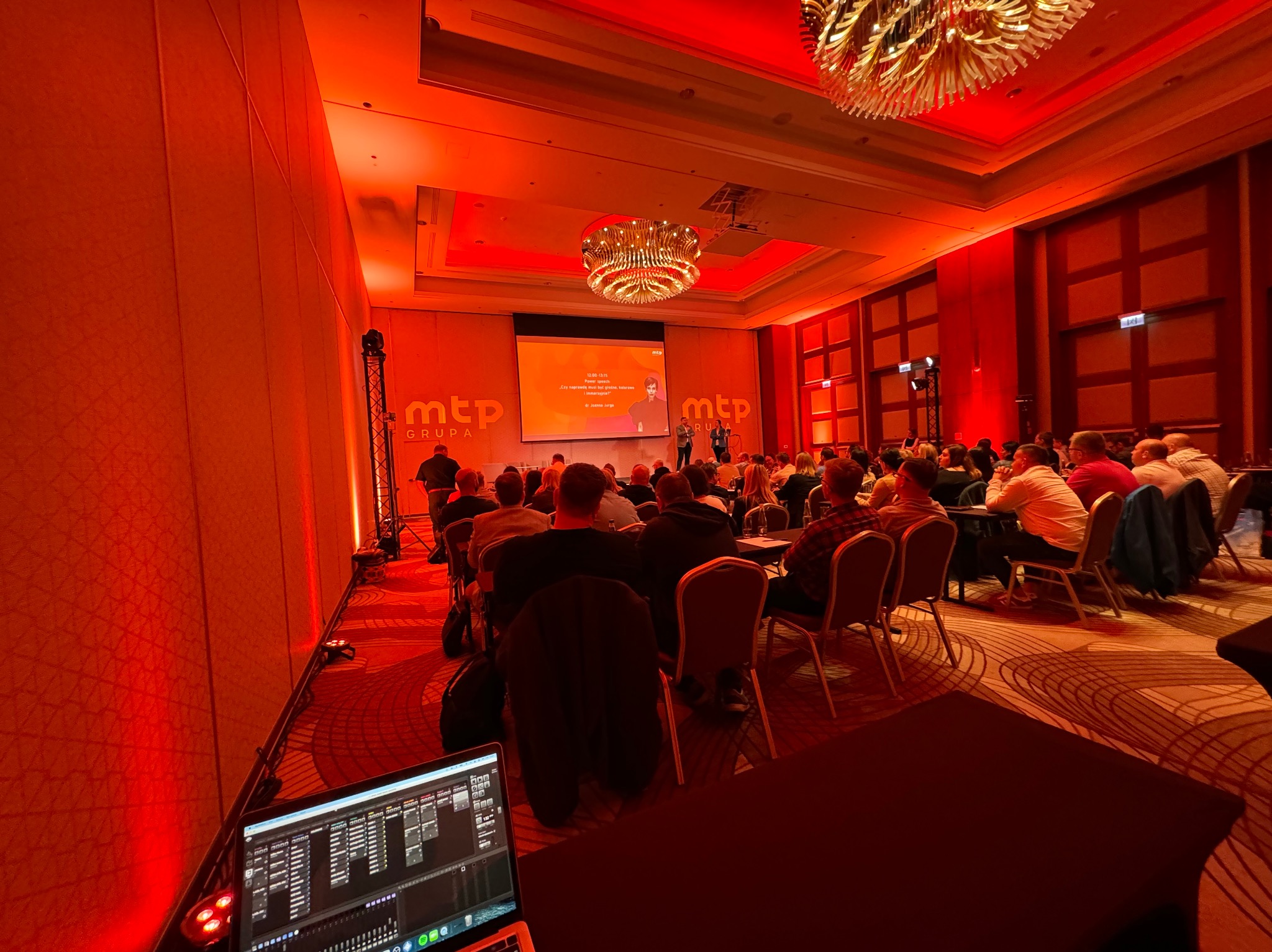 Sala konferencyjna oświetlona na czerwono, z publicznością siedzącą na krzesłach i prelegentami na scenie. Na pierwszym planie widoczny laptop z oprogramowaniem.