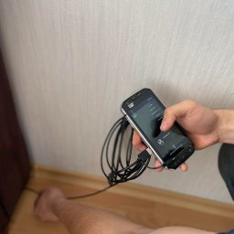 Cleanco Kacper Silecki - Osuszanie ściany przy podłodze za pomocą specjalistycznego urządzenia z podłączonymi kablami, trzymanego w dłoni. Widoczny fragment ściany i podłogi.