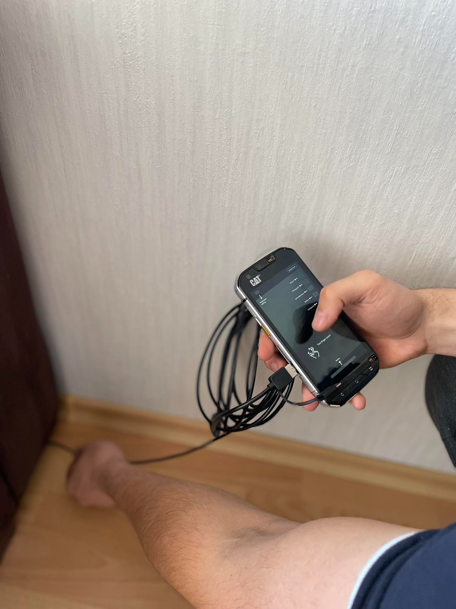 Osuszanie ściany przy podłodze za pomocą specjalistycznego urządzenia z podłączonymi kablami, trzymanego w dłoni. Widoczny fragment ściany i podłogi.