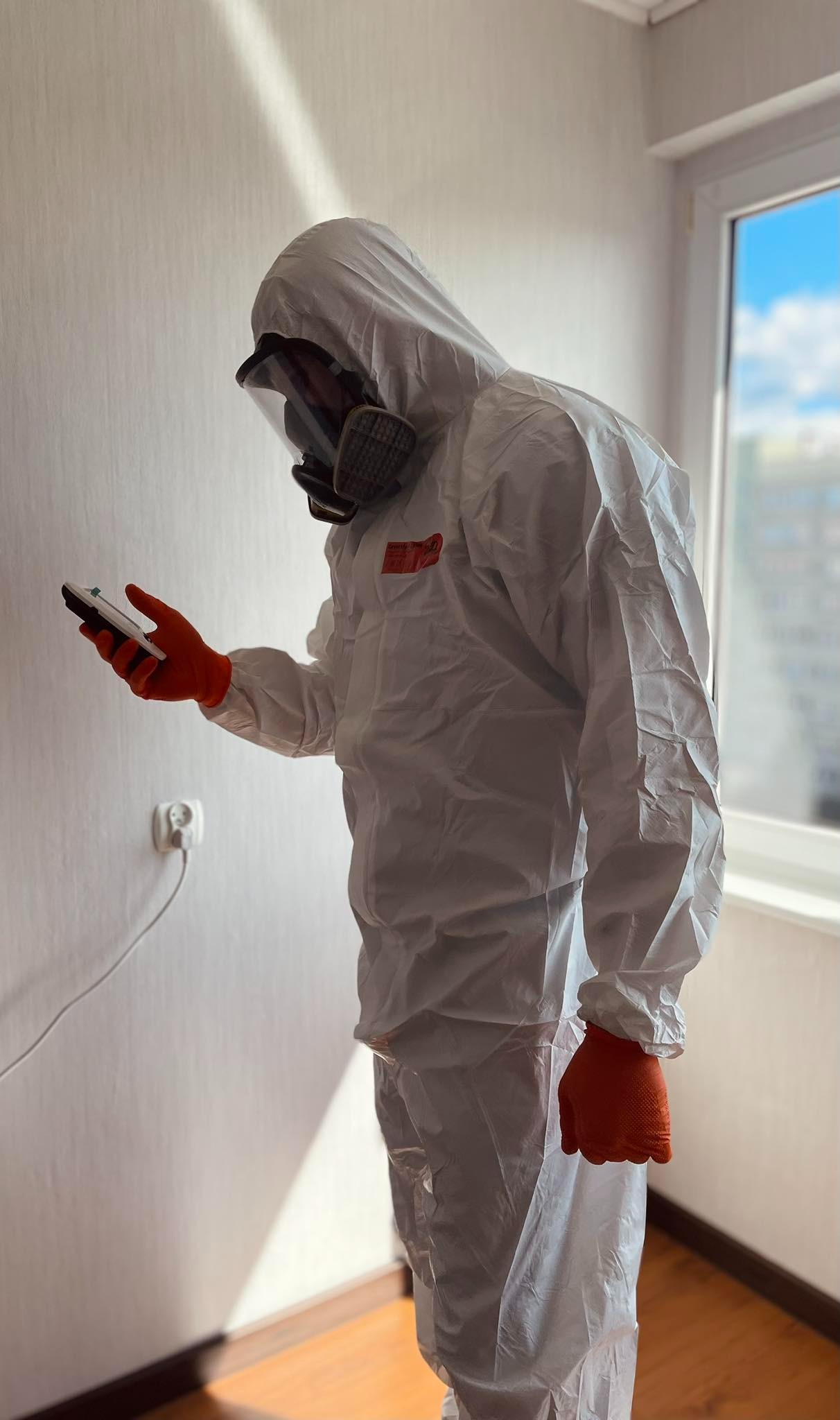 Osoba w kombinezonie ochronnym i masce sprawdza urządzeniem pomiarowym ścianę w jasnym pomieszczeniu z oknem. Widoczna wtyczka w gniazdku.