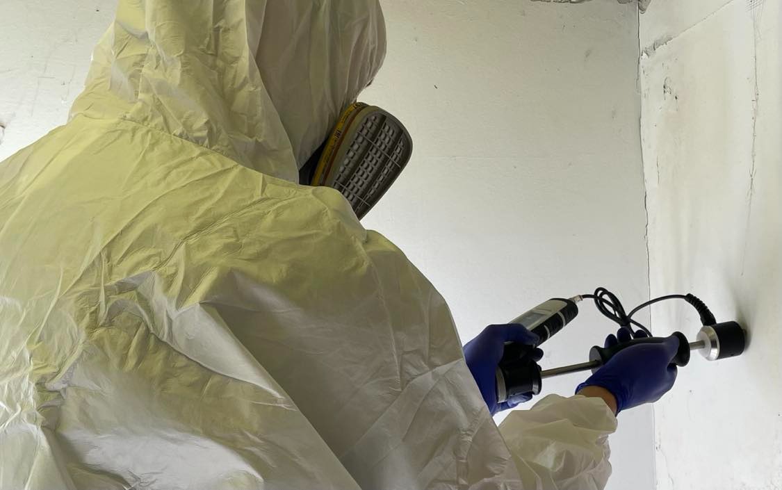 Pracownik w kombinezonie ochronnym mierzy wilgotność ściany za pomocą specjalistycznego urządzenia pomiarowego.