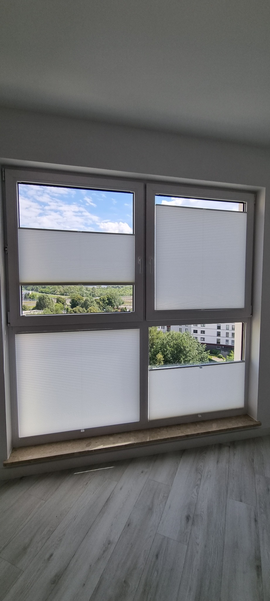 Białe plisy okienne zamontowane w oknie z widokiem na zieleń. Plisy częściowo zasłonięte, widać strukturę materiału. Nowoczesne rozwiązanie do regulacji światła.