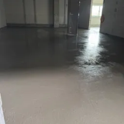 Świeżo wylana, gładka, jasnoszara powierzchnia w nowym pomieszczeniu z kolumnami. Widoczne ślady po narzędziach i refleksy świetlne. W tle ściany zabezpieczone folią.