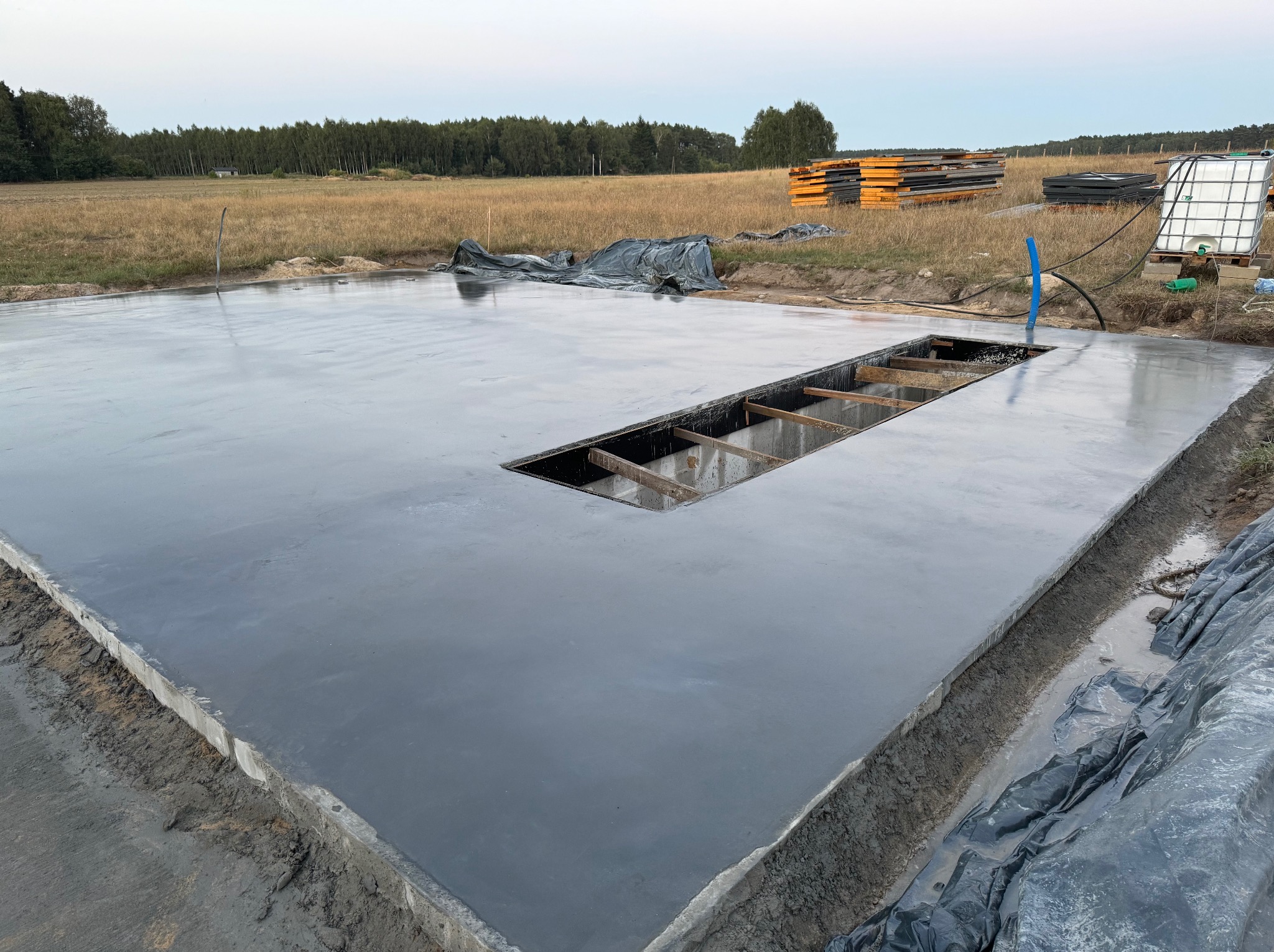 Świeżo wylana, gładka wylewka betonowa z otworem na instalacje, widok z góry, otoczenie placu budowy z elementami izolacji i zbiornikiem na wodę w tle.