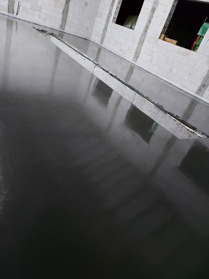 Świeżo wylana, ciemna posadzka przemysłowa, z widoczną listwą dylatacyjną w surowym wnętrzu budynku z białego bloczka.