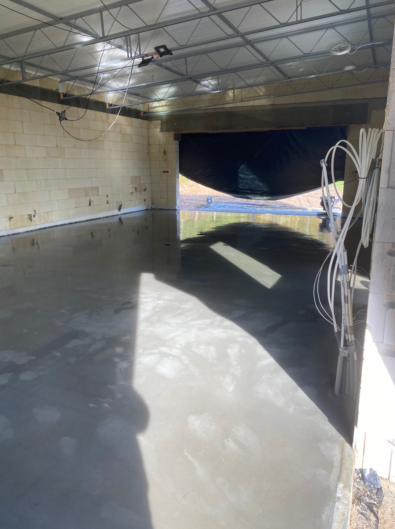 Świeża, gładka powierzchnia podłogi z wylewki w surowym wnętrzu hali, z widocznymi przewodami i blokami konstrukcyjnymi. W tle zarys otwartego wjazdu.