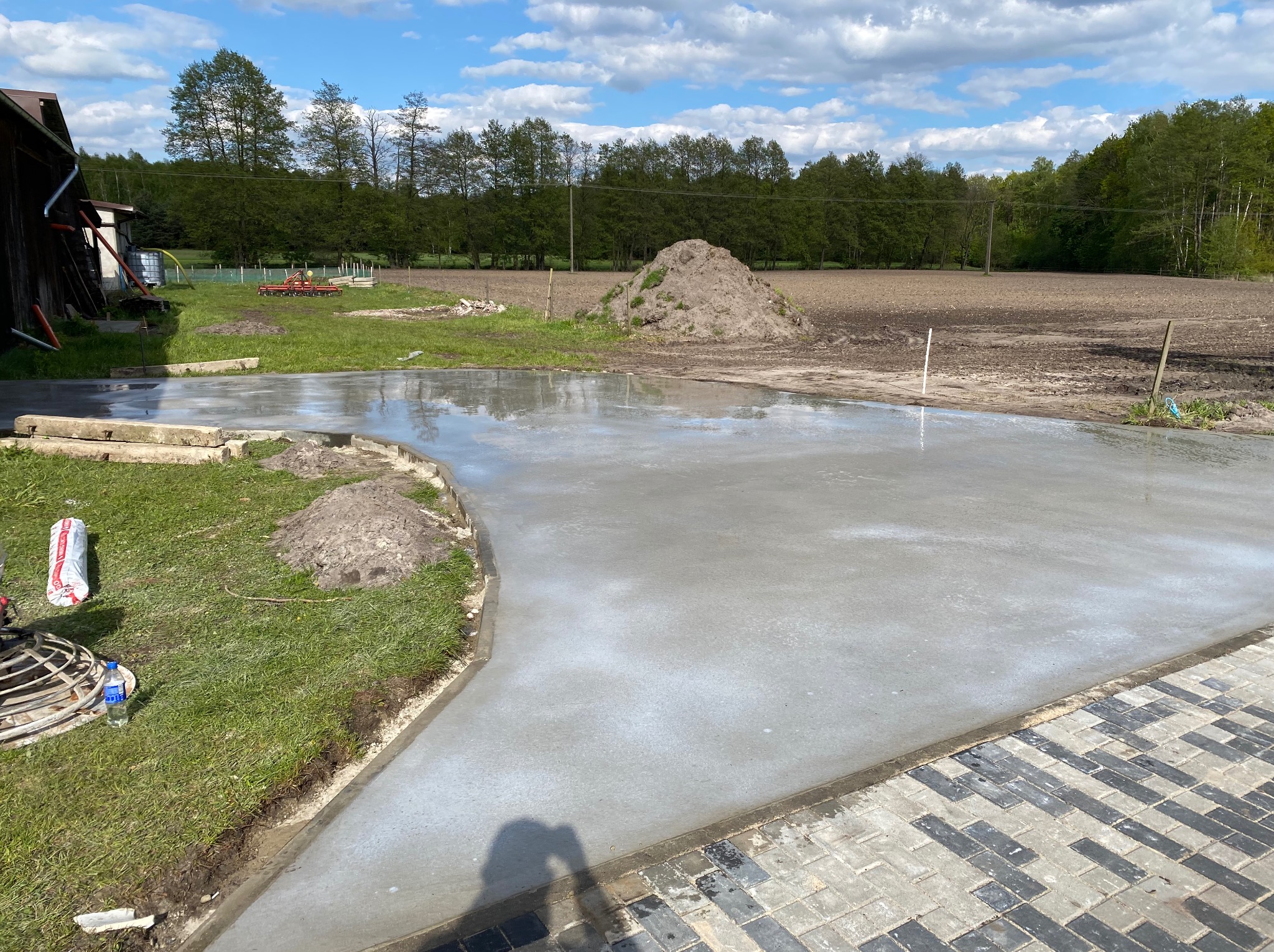 Świeżo wylana, gładka powierzchnia betonowa na zewnątrz, obok trawnik i kostka brukowa, w tle drzewa i błękitne niebo z chmurami.