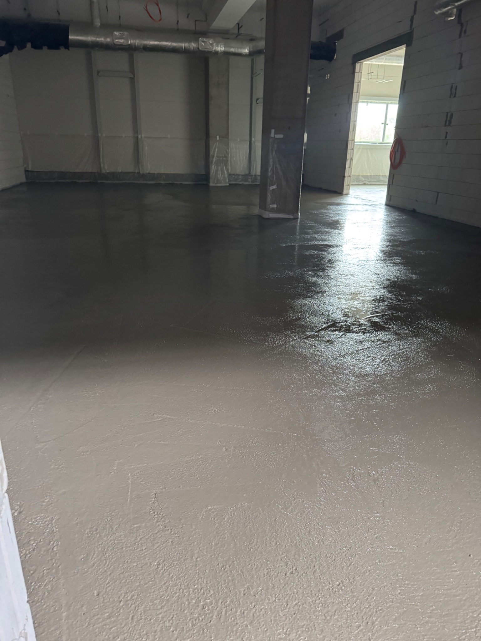 Świeżo wylana, gładka, jasnoszara powierzchnia w nowym pomieszczeniu z kolumnami. Widoczne ślady po narzędziach i refleksy świetlne. W tle ściany zabezpieczone folią.