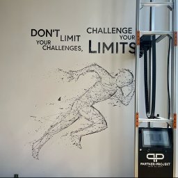 Partner-Projekt Druk Ścienny - Druk na ścianie w Krapkowicach: grafika biegacza i motywacyjne hasło wykonane za pomocą specjalistycznej drukarki pionowej, widocznej obok. Nowoczesny design.