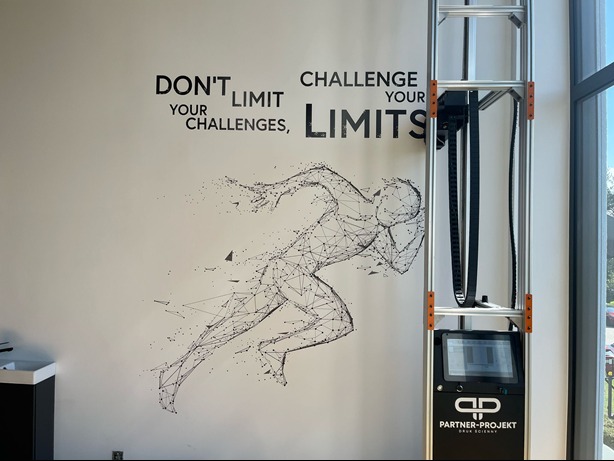 Druk na ścianie w Krapkowicach: grafika biegacza i motywacyjne hasło wykonane za pomocą specjalistycznej drukarki pionowej, widocznej obok. Nowoczesny design.