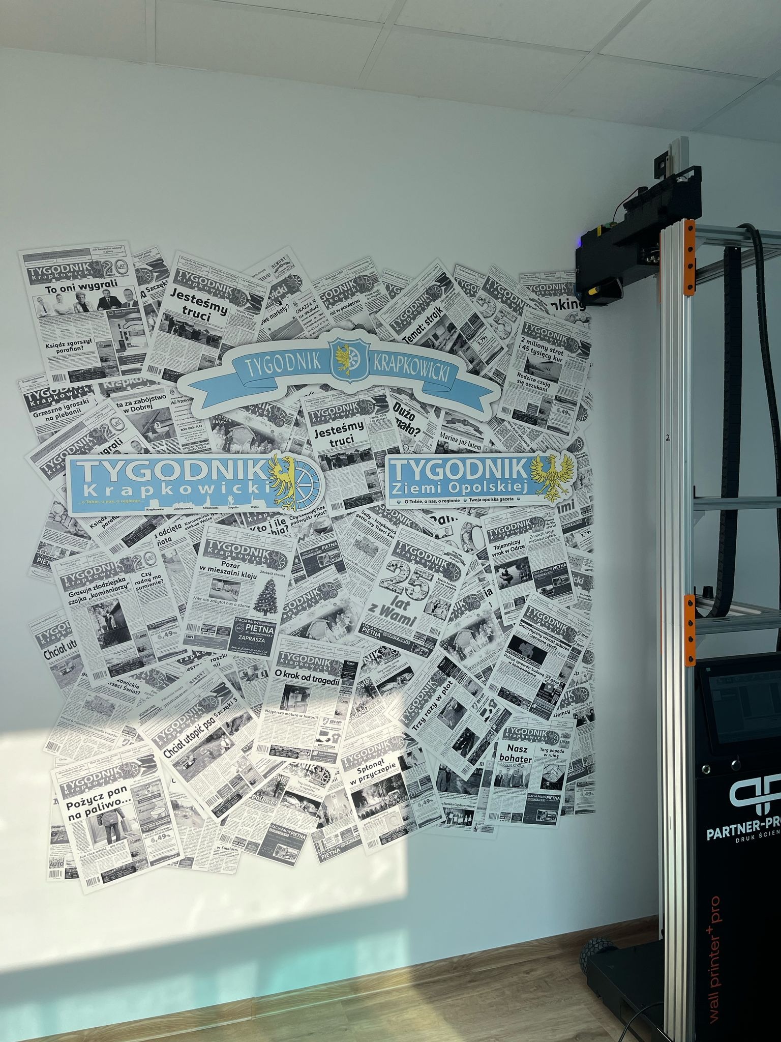 Druk na ścianie z użyciem plotera pionowego. Ściana pokryta gazetami 'Tygodnik Krapkowicki', na tle podłoga z paneli i biała ściana. Drukarka pionowa w trakcie pracy.