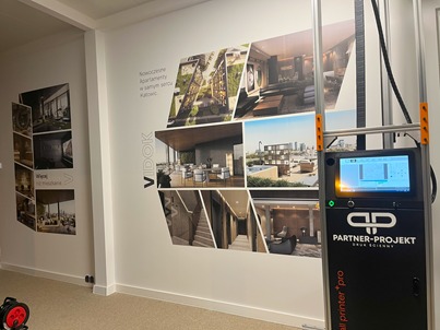 Drukarka ścienna aplikuje kolaż nowoczesnych apartamentów 'Vidok' w Katowicach na białą ścianę. Urządzenie PARTNER-PROJEKT w trakcie pracy.