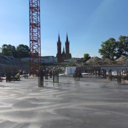 Betonoza - Świeżo wylana betonowa płyta fundamentowa z wystającymi prętami zbrojeniowymi na placu budowy. Pracownik w żółtej kamizelce obsługuje maszynę. W tle czerwony żuraw i kościół z dwiema wieżami.