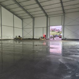 Betonoza - Duża hala z świeżo wylaną, mokrą, błyszczącą posadzką betonową, odbijającą światło i widok na domy z zewnątrz. W tle pracownicy z maszynami do wygładzania.