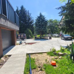 Betonoza - Szeroki, jasny podjazd z betonu, z widocznym wycięciem i niebieskim wężem, prowadzący do nowoczesnego domu z ciemną elewacją i garażem. W tle zieleń.