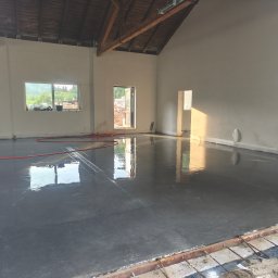 Betonoza - Lśniąca, świeżo wylana posadzka betonowa w przestronnym pomieszczeniu z wysokim sufitem, drewnianymi belkami i odbiciami światła dziennego, z czerwoną rurą.