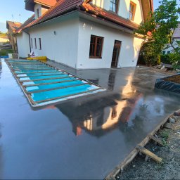Betonoza - Świeżo wylana, mokra płyta betonowa obok domu, z prostokątnym otworem zabezpieczonym drewnianymi listwami i niebieską izolacją. Na błyszczącej powierzchni odbija się fasada budynku.