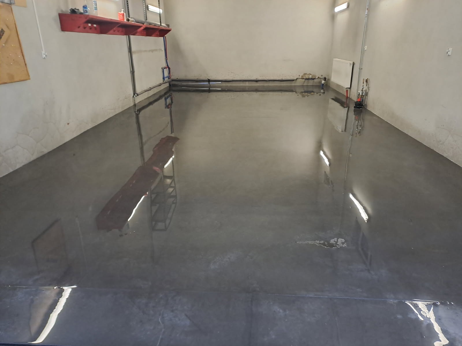 Błyszcząca, szara posadzka przemysłowa w jasnym pomieszczeniu, z widocznymi instalacjami na ścianach i odbiciami światła. Powierzchnia gładka, z drobnymi niedoskonałościami.
