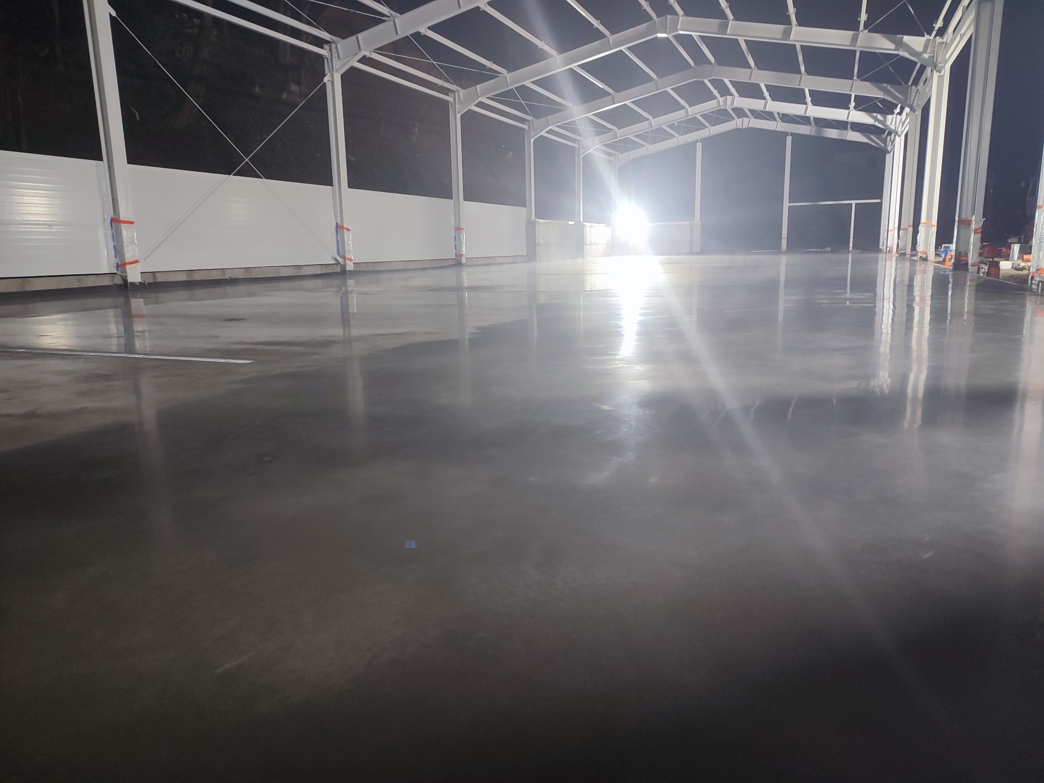 Wypolerowana, szara posadzka przemysłowa w hali z metalową konstrukcją. Świeżo wylana, gładka powierzchnia odbija światło. Widoczne białe ściany i słupy.