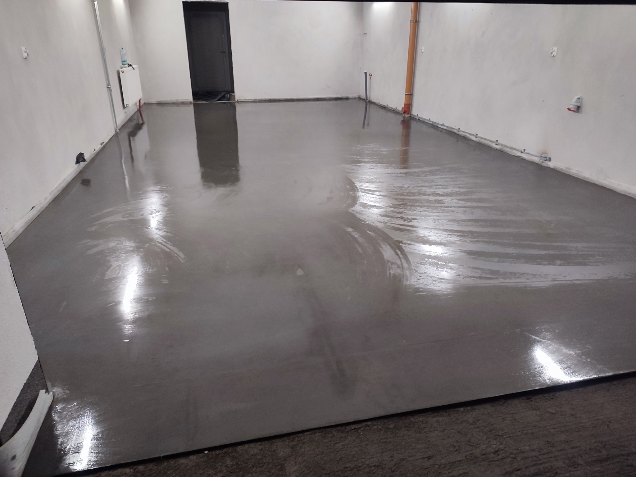 Świeżo wylana, gładka posadzka przemysłowa w jasnym pomieszczeniu. Widoczne ślady po rozprowadzaniu masy, rury i elementy instalacji przy ścianach.