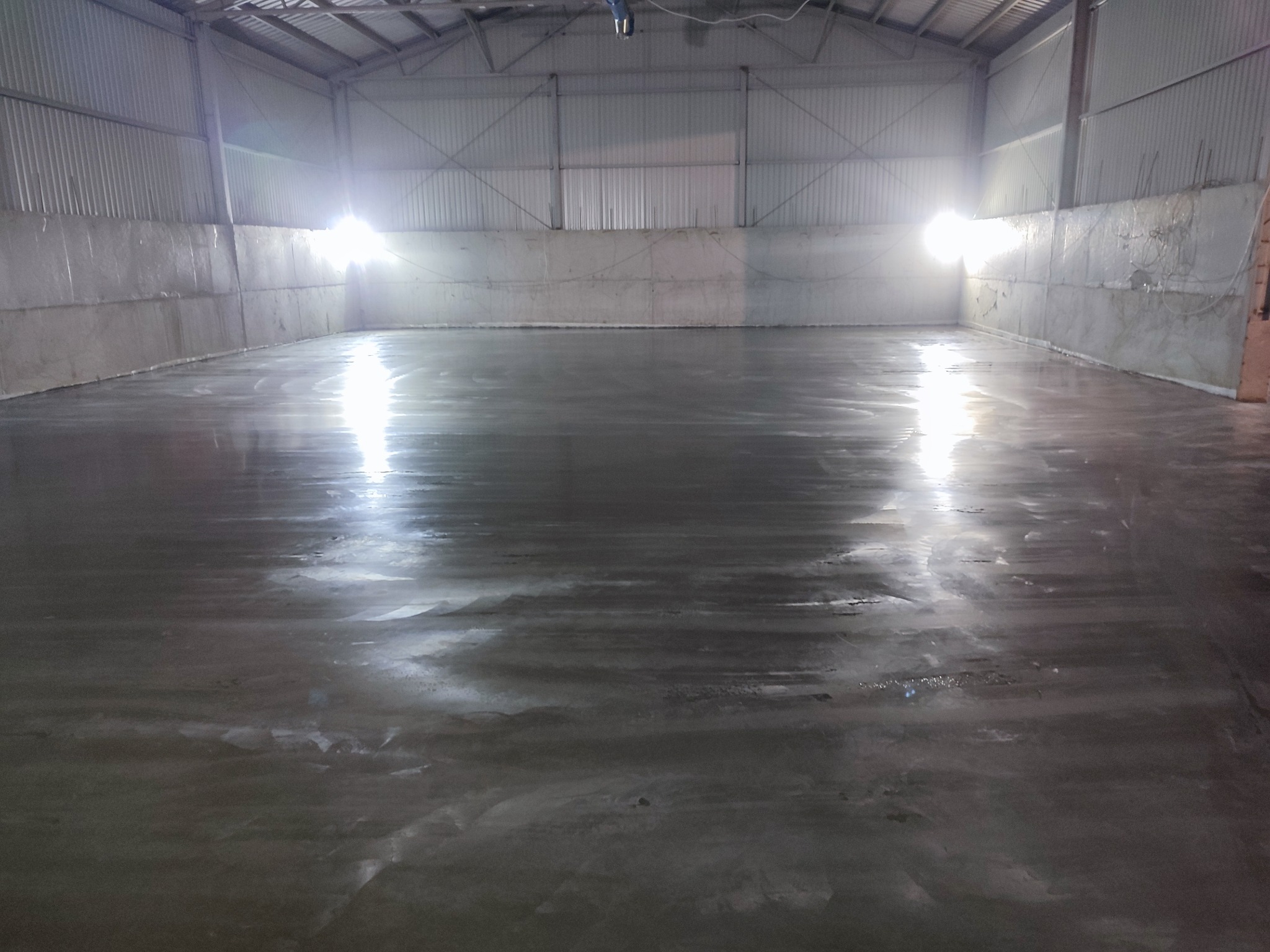 Gładka, mokra posadzka przemysłowa w hali z blachy falistej, odbijająca światło. Ściany częściowo wykończone, surowy beton. Widok na całą powierzchnię.