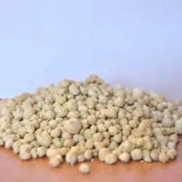 Jasnobrązowe granulki nawozu ułożone w małą kupkę na gładkiej powierzchni, z rozmytym, jasnym tłem. Widoczne pojedyncze granulki poza główną masą.