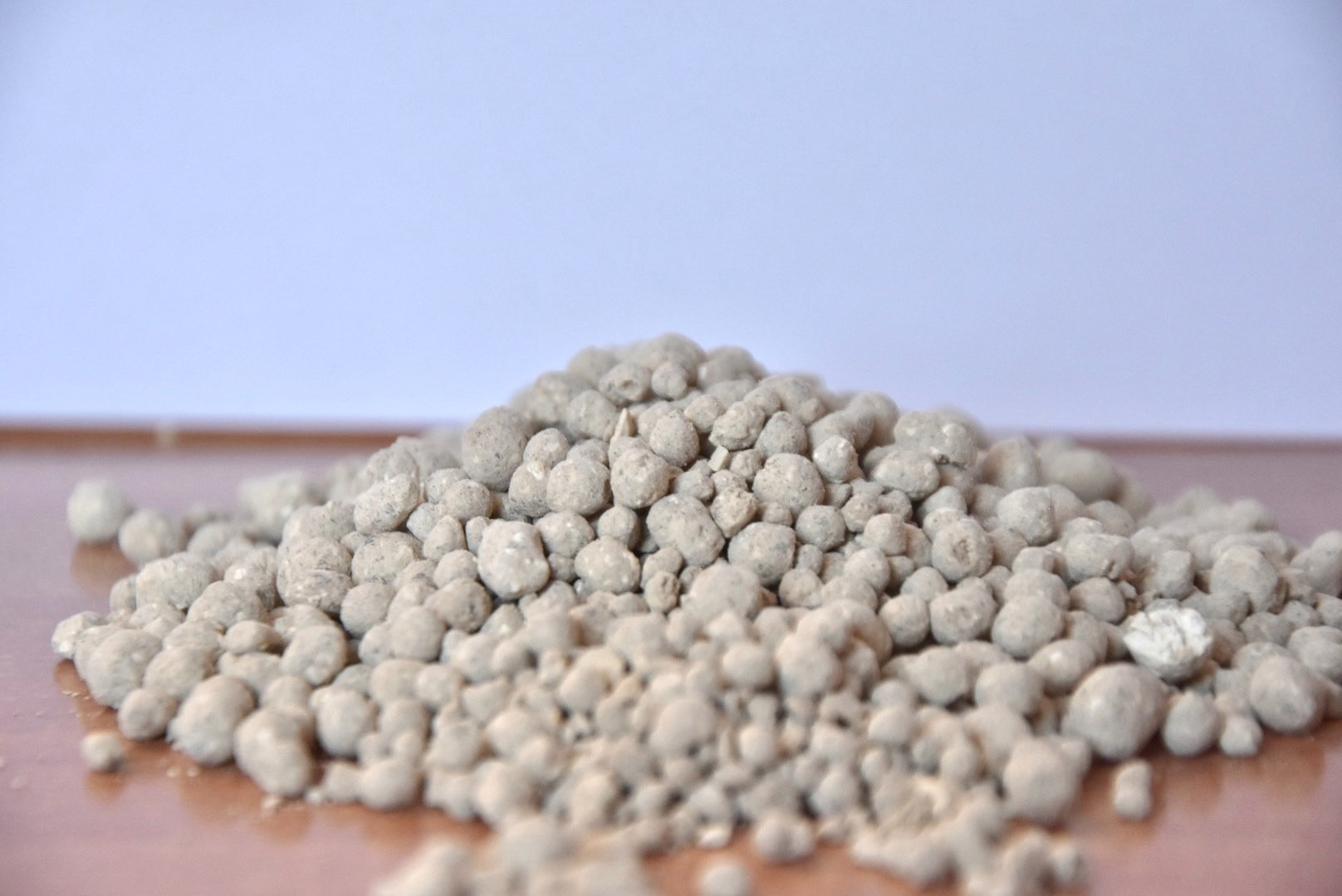 Kopuła szaro-białych granulek nawozu na gładkiej, brązowej powierzchni, z rozmytym, jasnym tłem. Granulki o nieregularnych kształtach i porowatej strukturze.