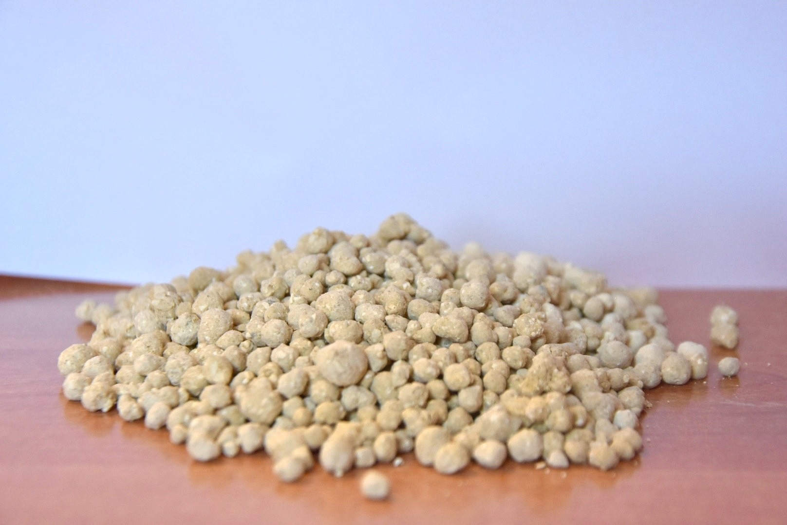 Jasnobrązowe granulki nawozu ułożone w małą kupkę na gładkiej powierzchni, z rozmytym, jasnym tłem. Widoczne pojedyncze granulki poza główną masą.