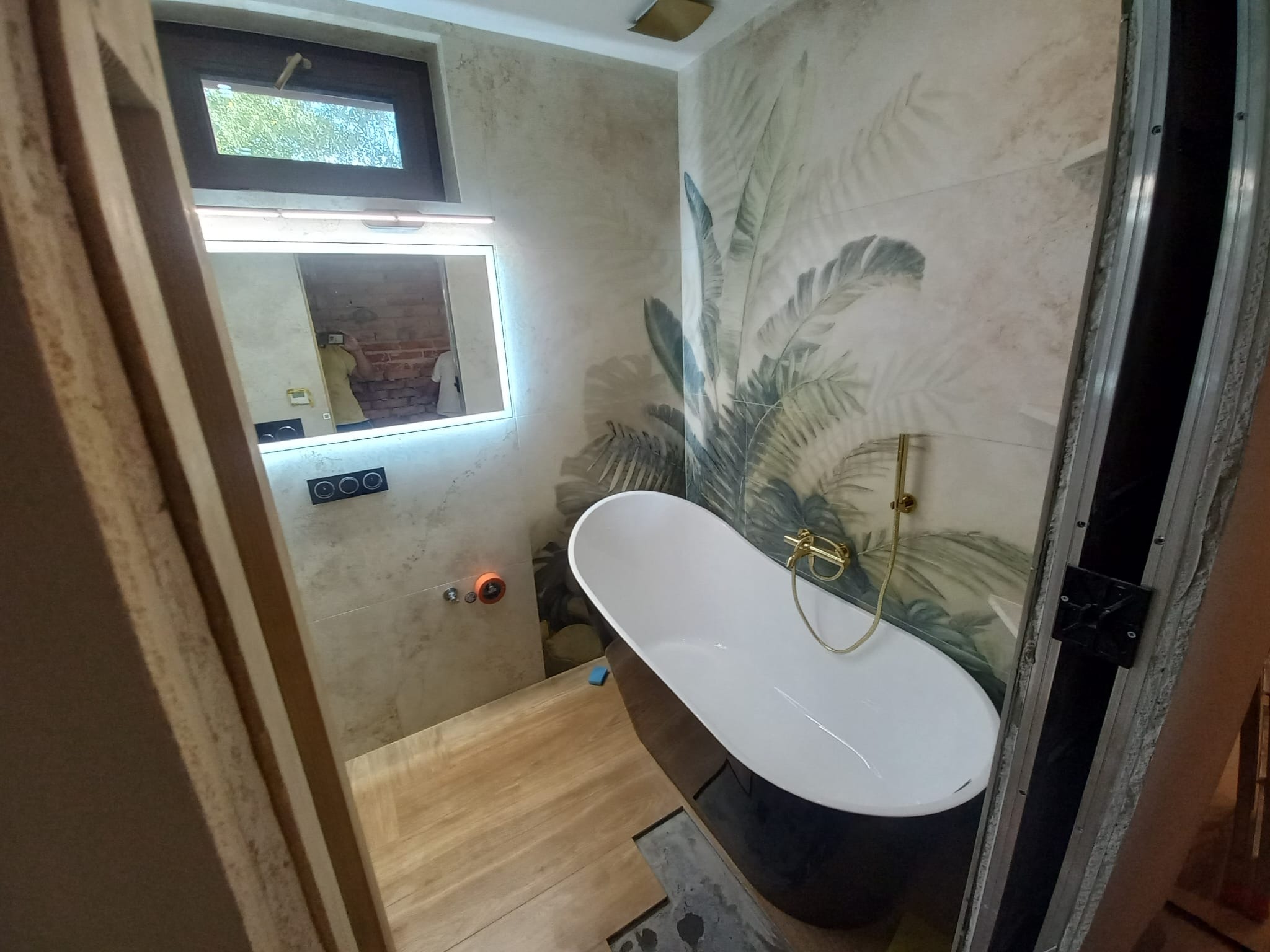 Nowoczesna łazienka w trakcie wykańczania: wolnostojąca wanna na drewnopodobnej podłodze, płytki z motywem roślinnym, lustro z podświetleniem LED. Widoczne detale montażowe.