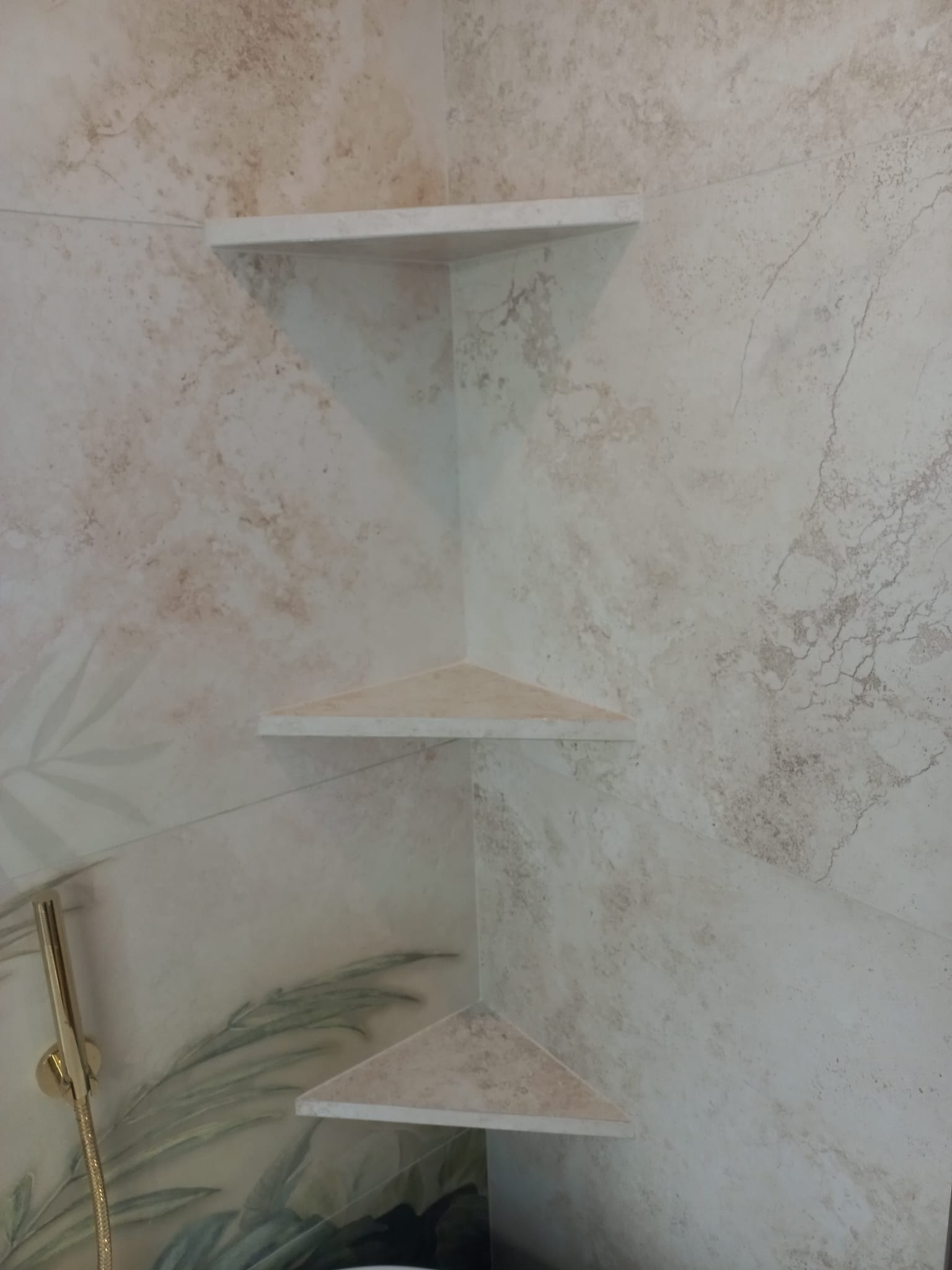 Wykończona kabina prysznicowa z trzema narożnymi, trójkątnymi półkami z płytek w odcieniach beżu i brązu, z motywem roślinnym na ścianie i złotym prysznicem.