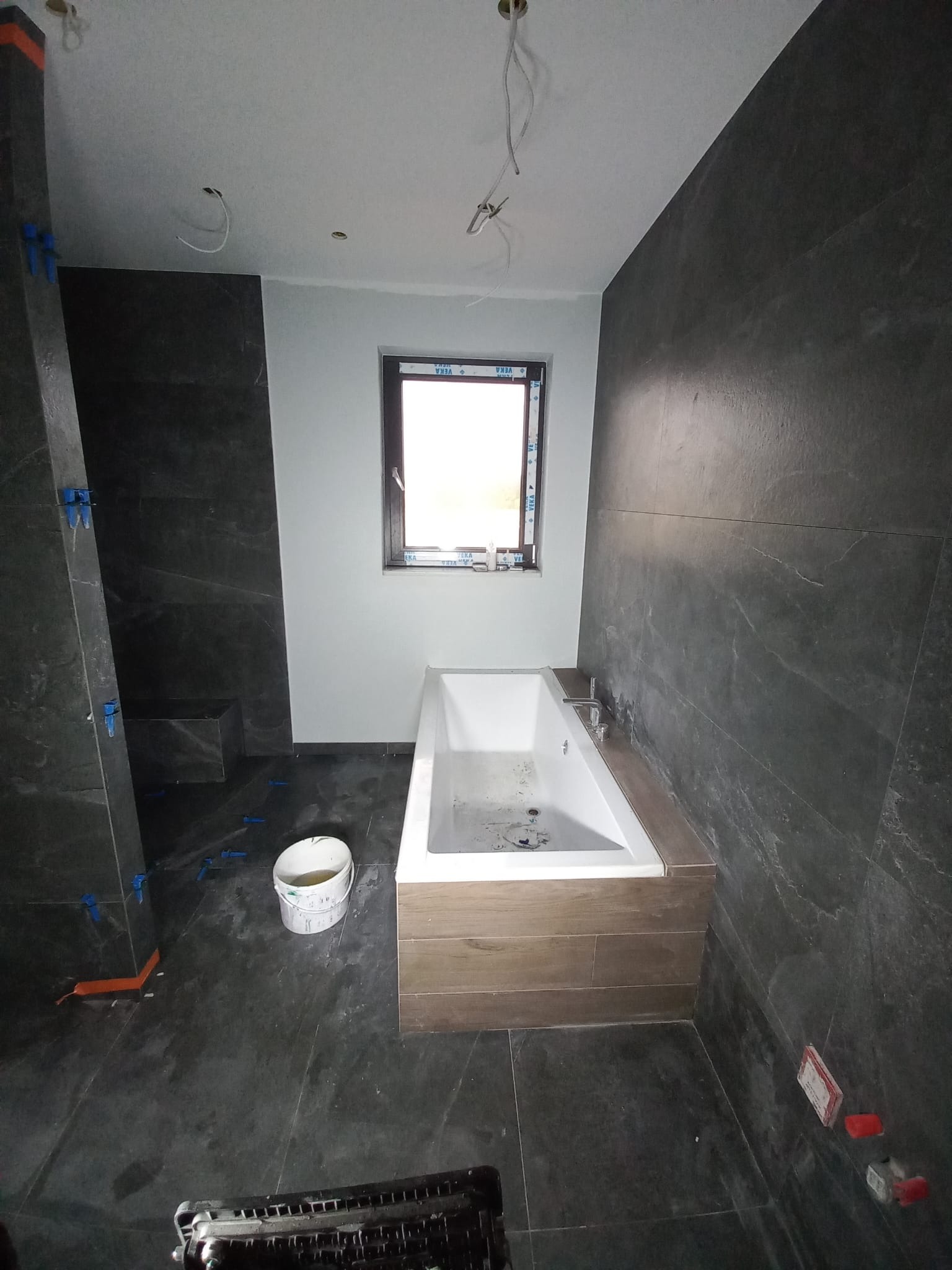 Wykończona łazienka w ciemnych płytkach z wanną w zabudowie. Widoczne narzędzia glazurnicze, płytki z fugą. Układanie płytek w trakcie realizacji.