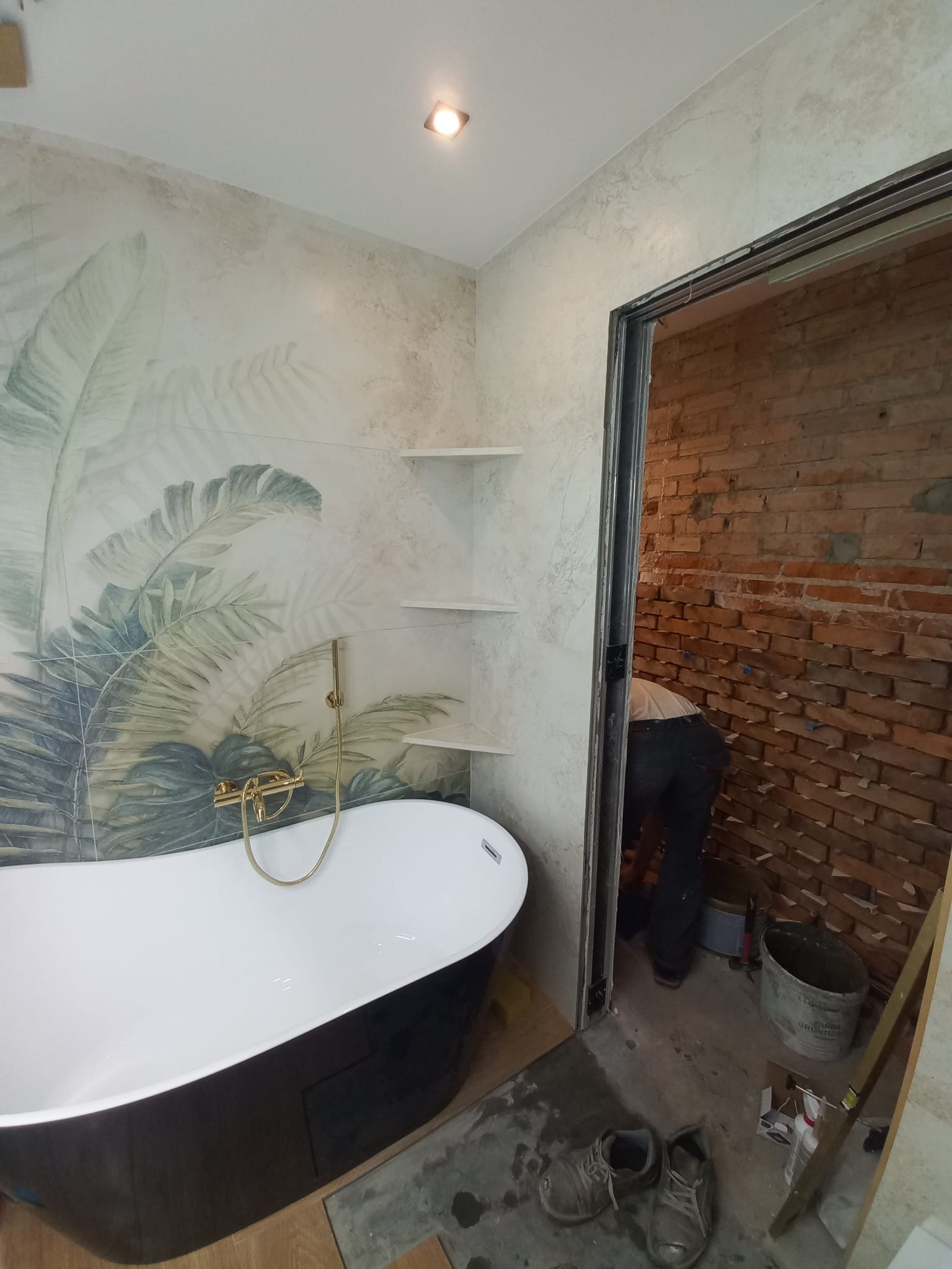 Łazienka w trakcie prac: wolnostojąca wanna z baterią w kolorze złota, tropikalna tapeta i półki narożne. W tle mur z cegły przed wykończeniem.