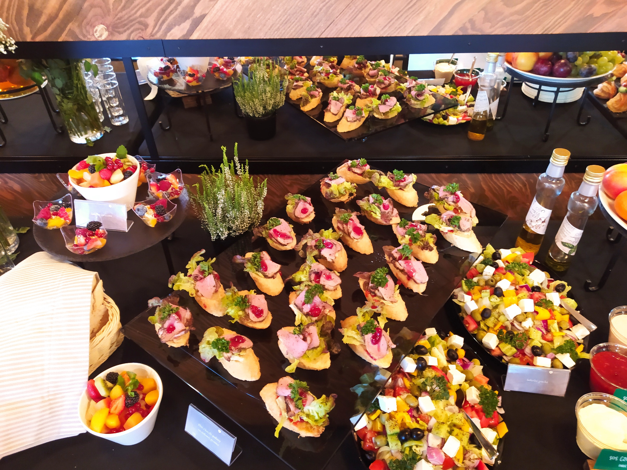 Elegancki stół cateringowy z przekąskami: bruschetta z rostbefem, sałatka grecka, owoce. Czarny blat, drewniane tło, dekoracje roślinne. Profesjonalny catering.