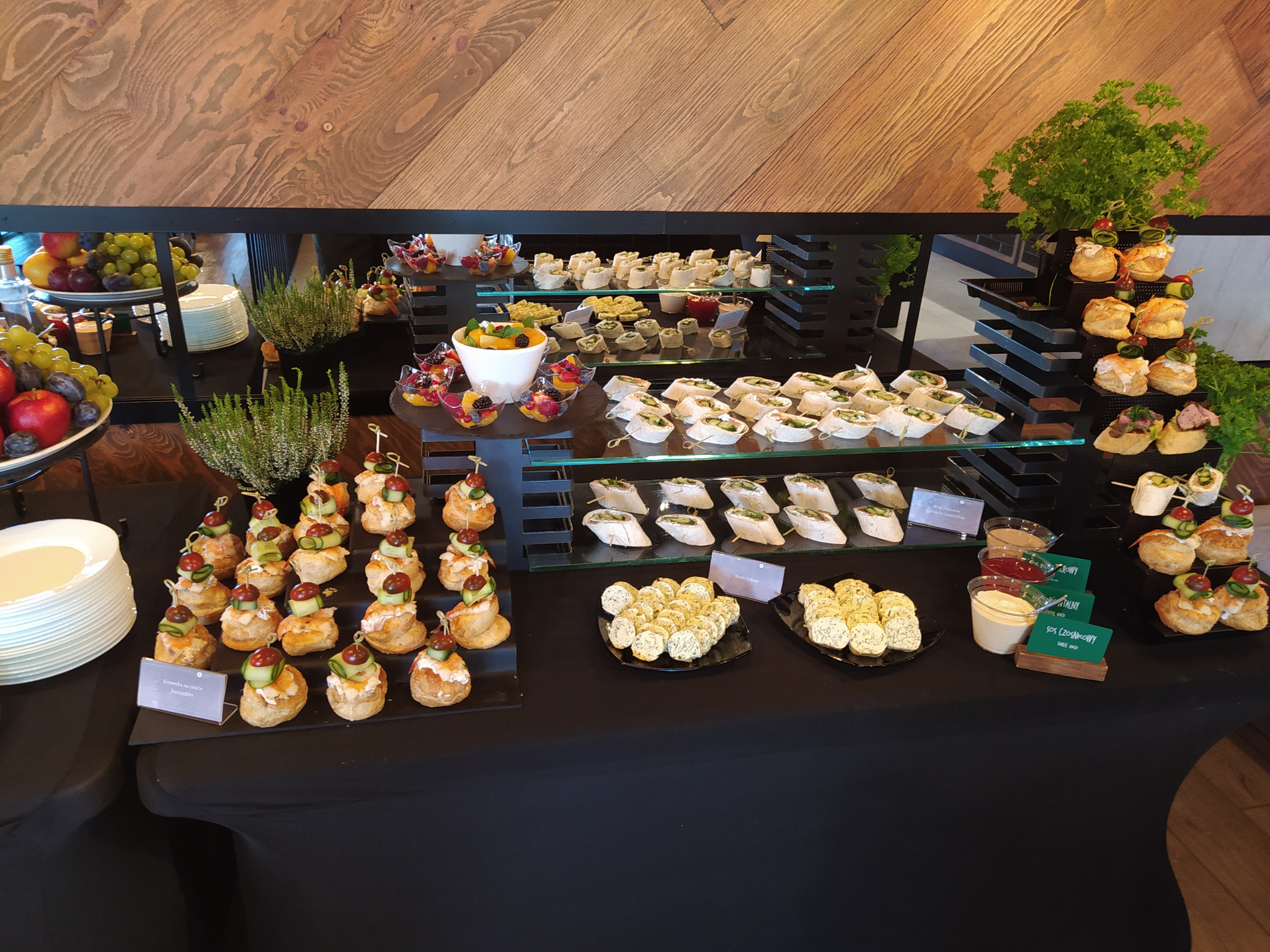 Bogato zastawiony stół cateringowy z różnorodnymi przekąskami: kanapeczki, tartinki, sałatki owocowe i sosy. Elegancki wystrój i dbałość o detale.