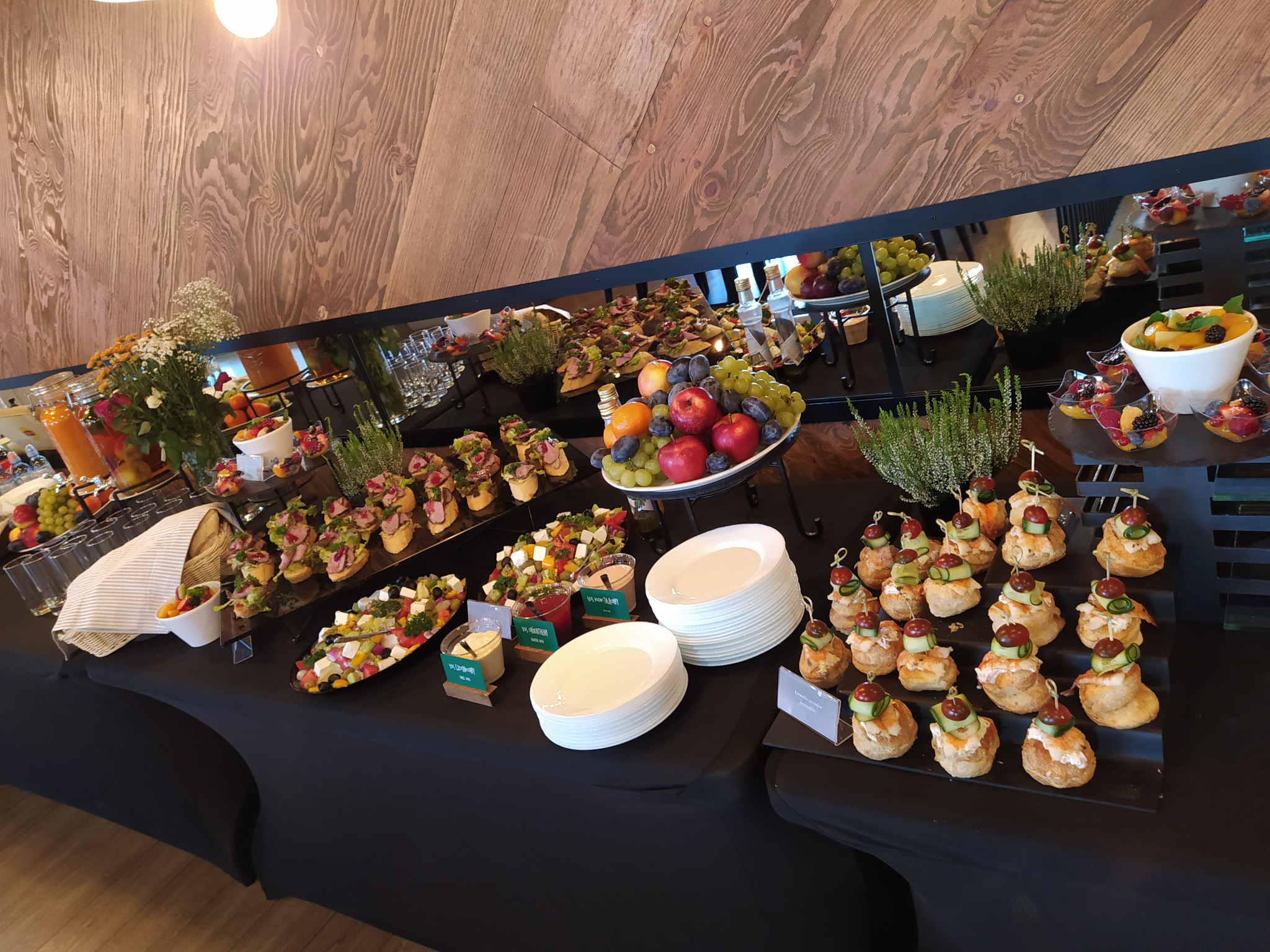 Stół cateringowy z różnorodnymi przekąskami: kanapeczki, sałatki owocowe, talerze z owocami. Czarny obrus, drewniane tło. Elegancki bufet.