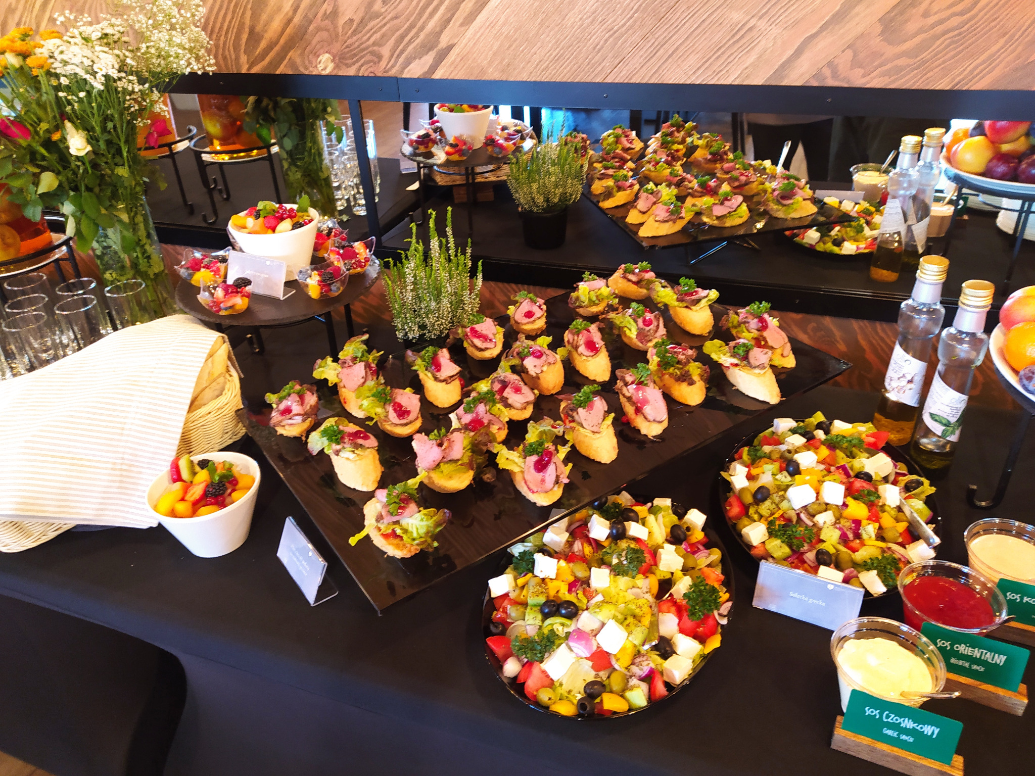 Stół cateringowy z przekąskami: kanapki z mięsem, sałatki, owoce, sosy. Dekoracje kwiatowe i ziołowe. Elegancki bufet na imprezę firmową lub świąteczną.