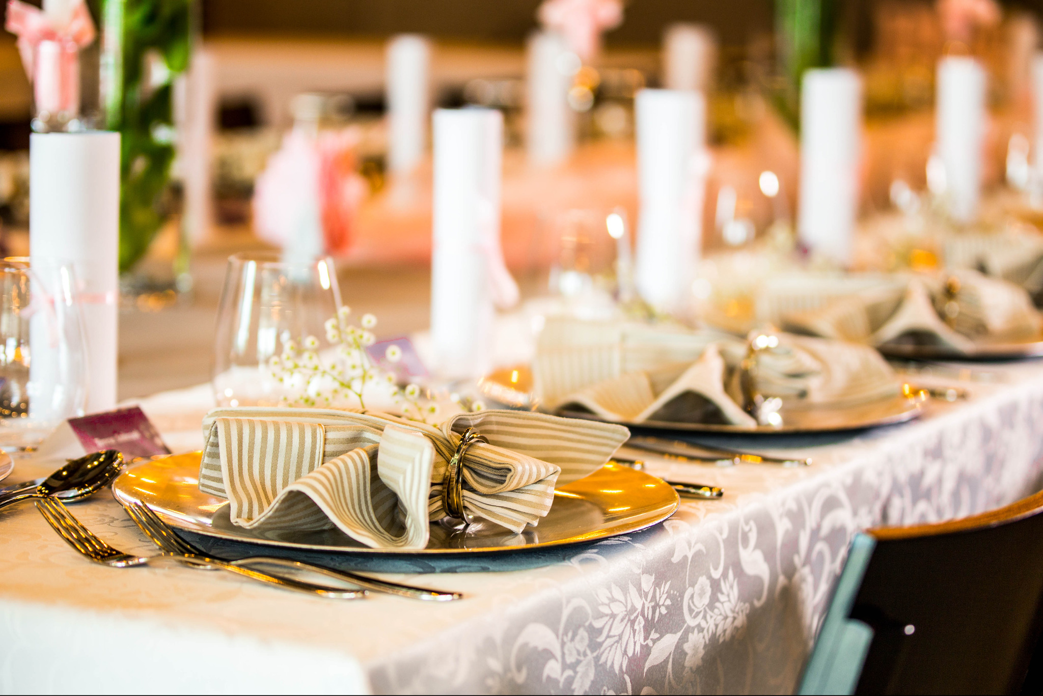 Uroczysty stół z elegancką zastawą: złote talerze, beżowe serwetki w paski z pierścieniami, sztućce. W tle świece, kwiaty i dekoracje w odcieniach bieli i różu.