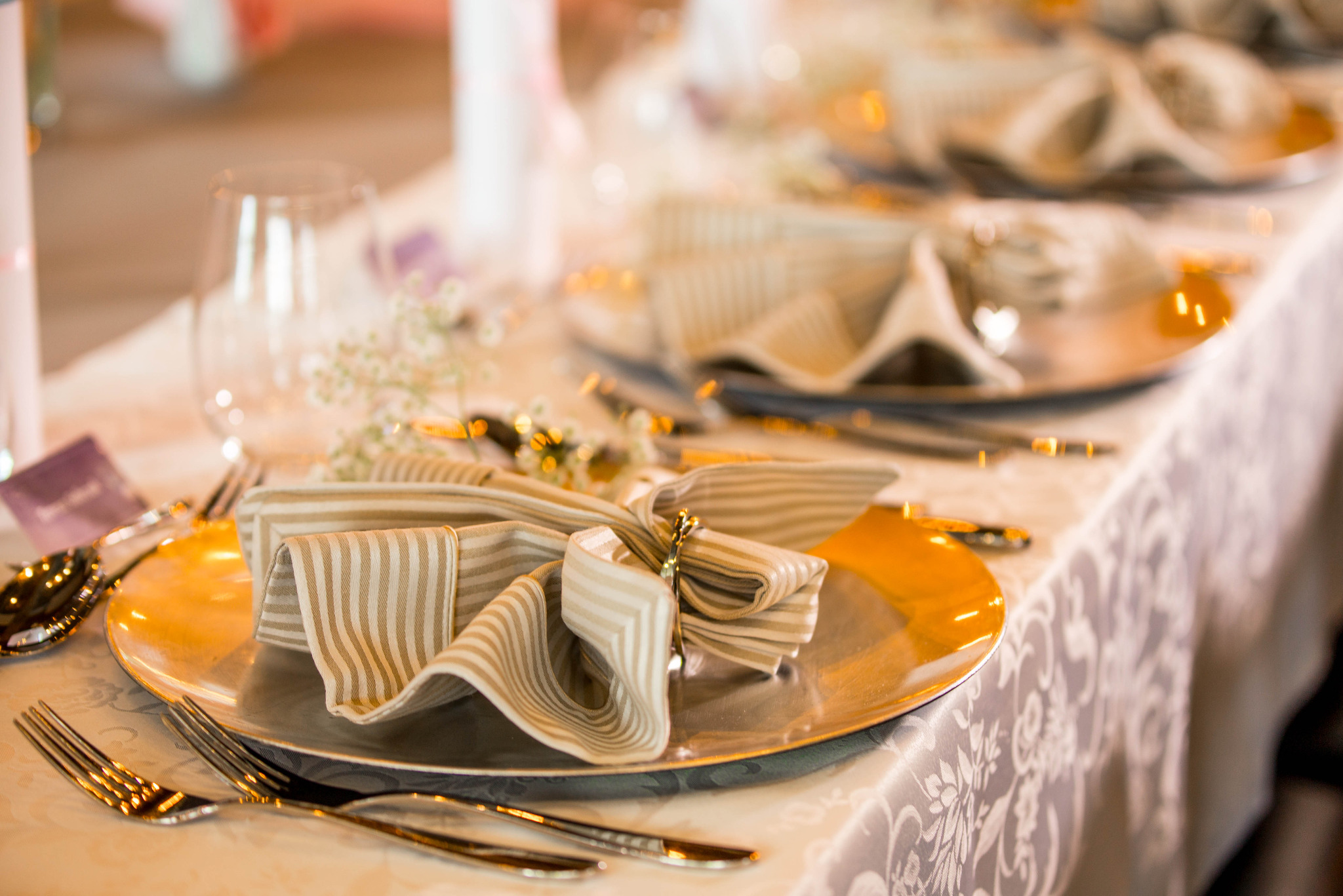 Elegancko nakryty stół: srebrne talerze, beżowe serwetki w paski, złote sztućce i delikatne kwiaty. Idealne na wykwintne przyjęcie.