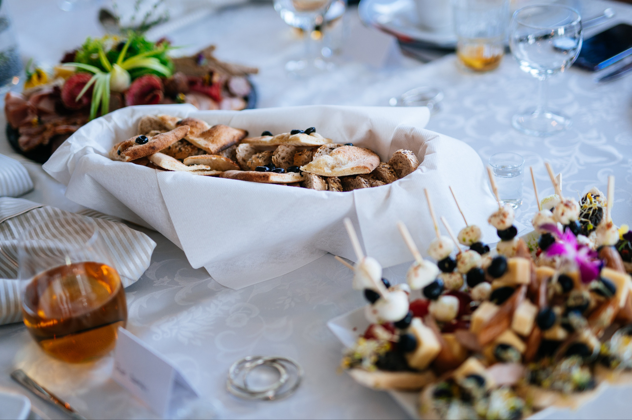 Elegancki stół z przekąskami: pieczywo w koszyku z serwetką, koreczki z serem i winogronami, wędliny i sery. Całość w jasnej tonacji, idealna na firmowe przyjęcie.